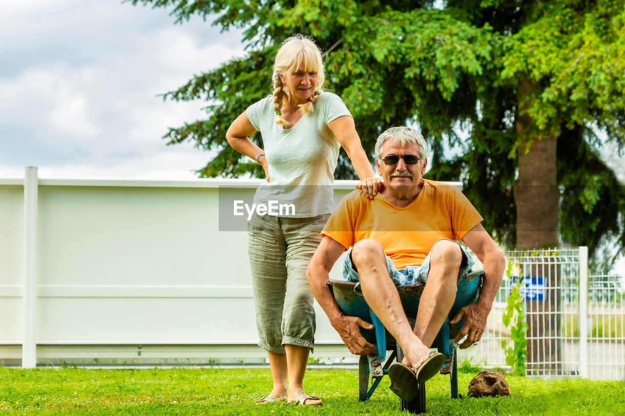 Senior man sitting on wheelbarrow while standing with wife at yard