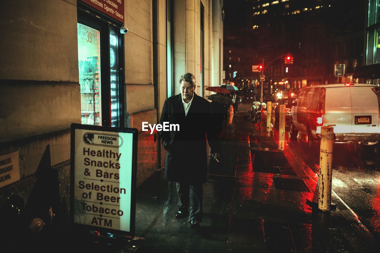 MAN STANDING ON ILLUMINATED CITY STREET AT NIGHT