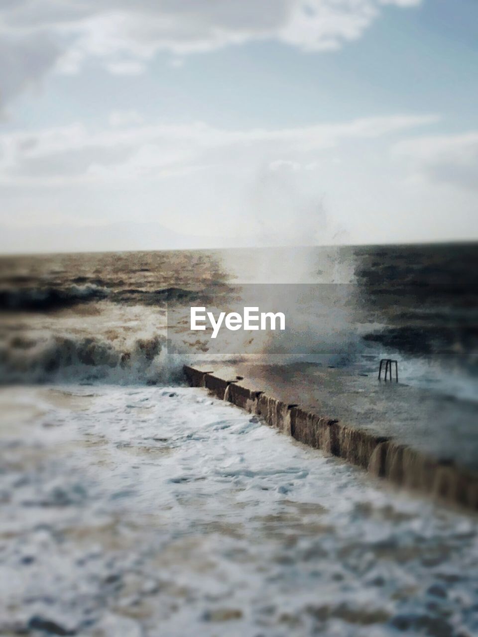 Tilt-shift image of wave splashing on pier against sky