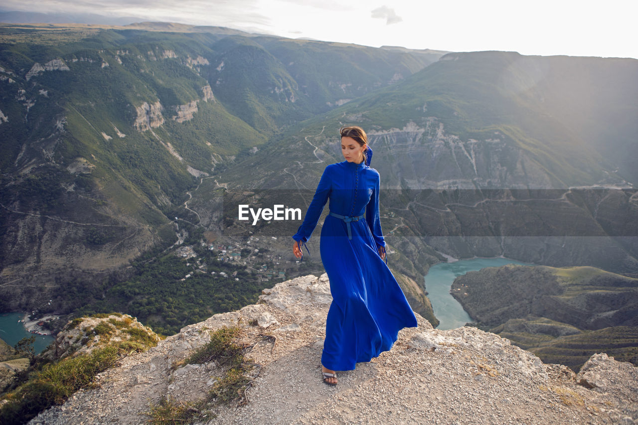 Brunette woman in a blue long dress stands on the edge of the sulak canyon in the evening at sunset