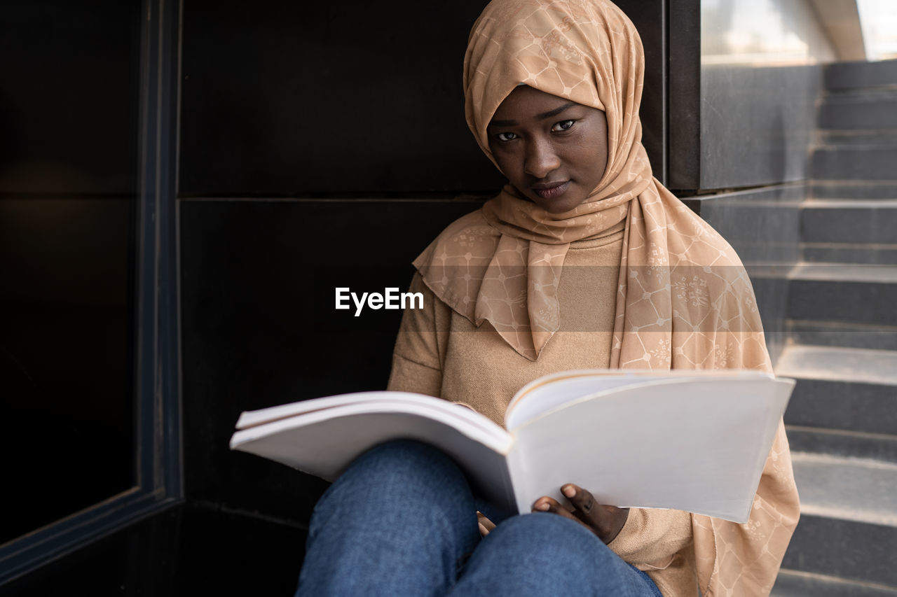 Crop concentrated young african american muslim female student in traditional headscarf sitting on street and reading book while preparing for exams