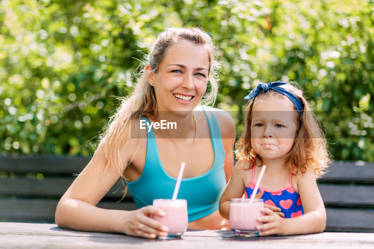 A beautiful girl and her little daughter drink a refreshing smoothie while sitting on a bench 