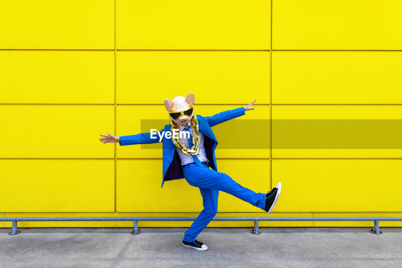 Man wearing vibrant blue suit, pig mask and large golden chain standing on one leg in front of yellow wall