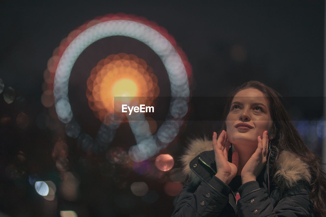 Thoughtful young woman standing against illuminated ferris wheel at night