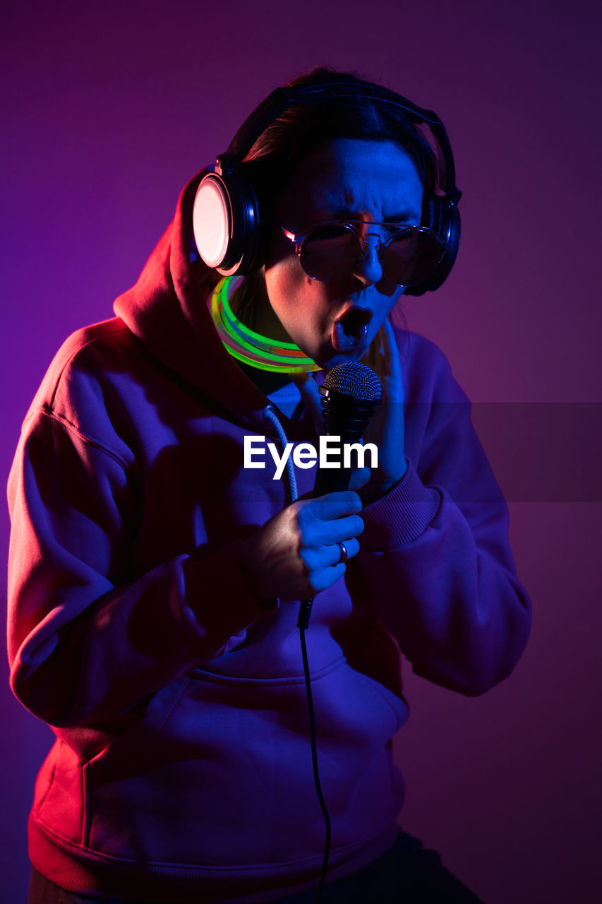 Cyberpunk woman in hooded hoodie and sunglasses dances against  wall with neon sticks and headphones