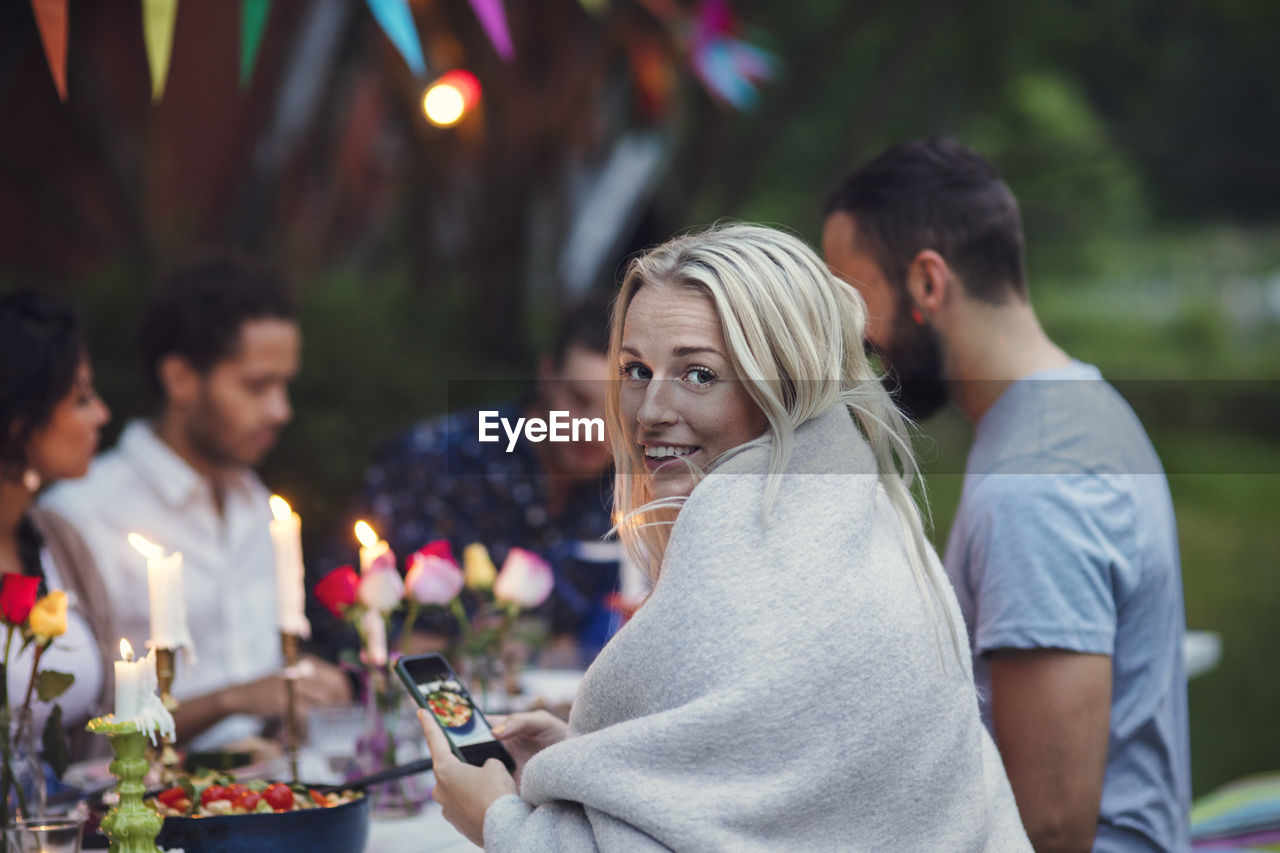 Portrait of smiling woman photographing food while sitting with friends at garden party