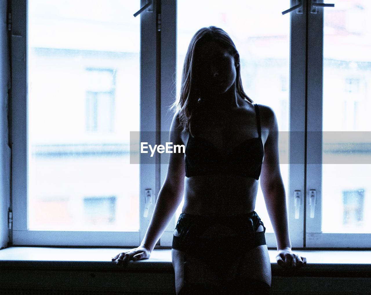 REAR VIEW OF YOUNG WOMAN STANDING AGAINST WINDOW