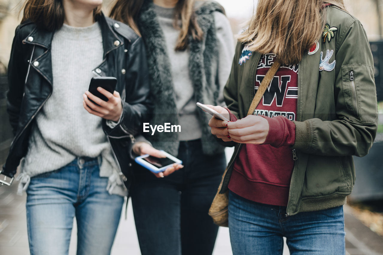 Midsection of female friends using mobile phone while walking in city