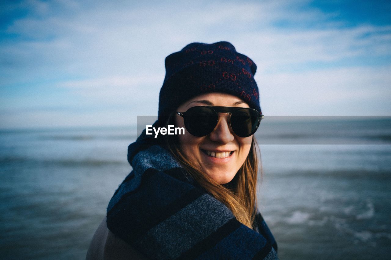 Close-up portrait of young woman wearing sunglasses while standing at beach against sky