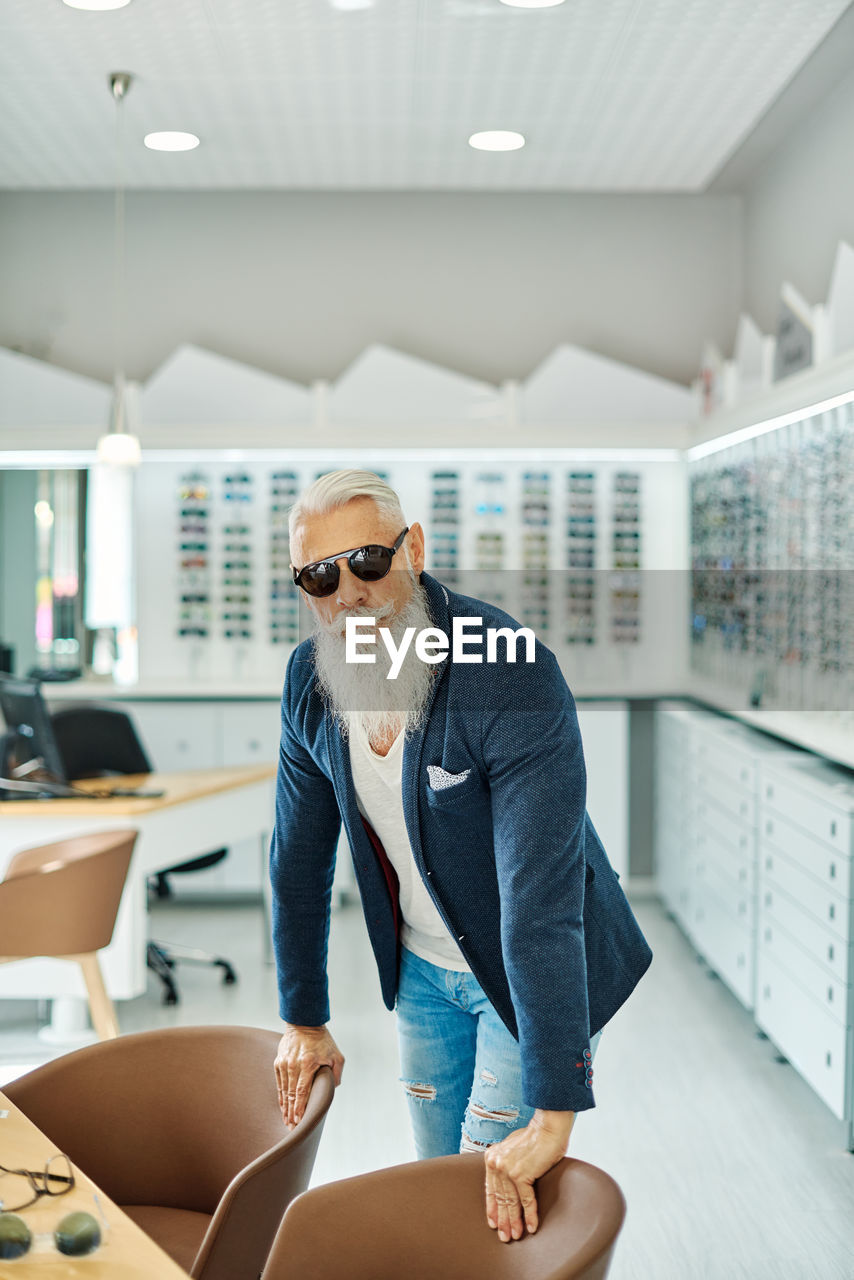 Self confident elderly male in trendy sunglasses and outfit standing in optical store and looking at camera