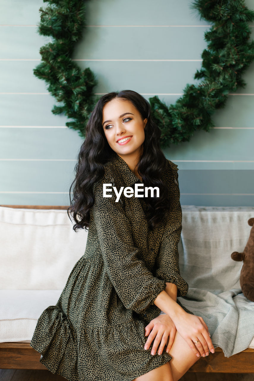 A young woman in a dress sitting on a light sofa against a blue wall with a christmas wreath 