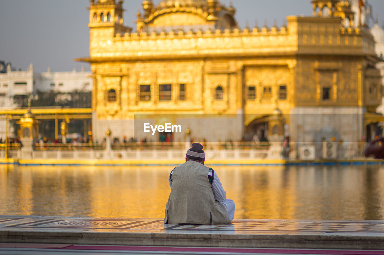 Rear view of man looking at golden temple while sitting on retaining wall by pond