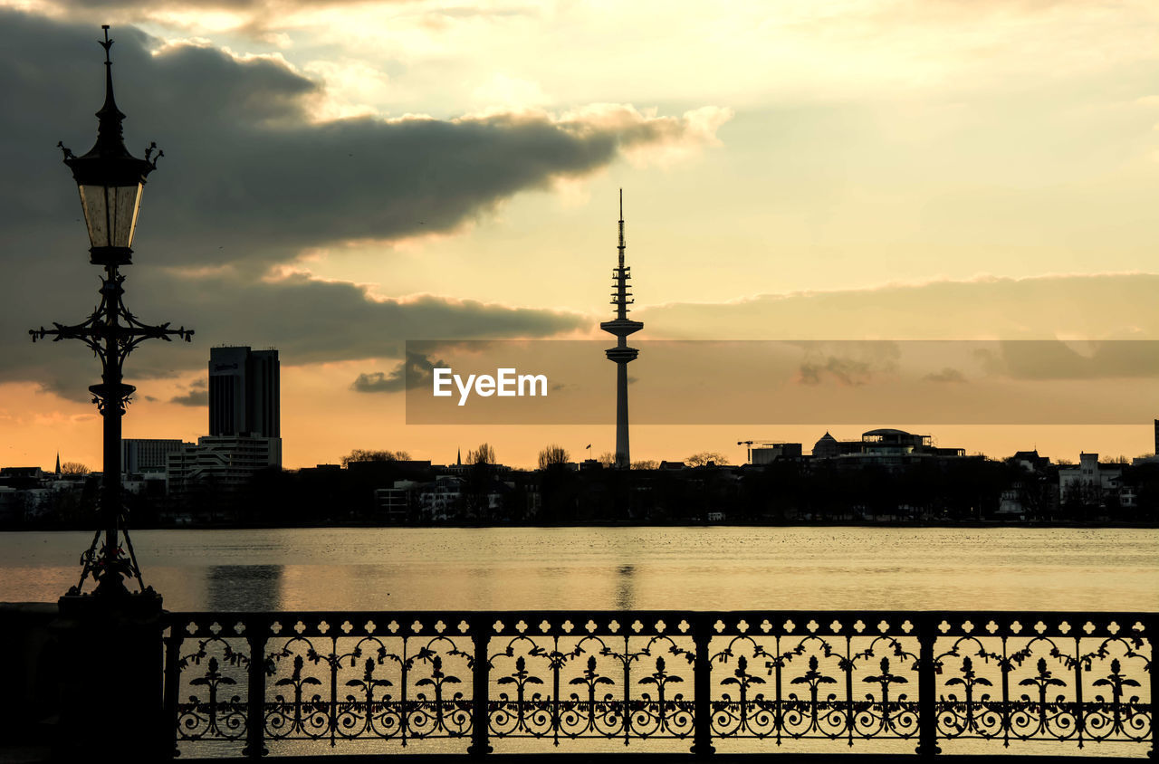 Silhouette heinrich-hertz-turm and cityscape by river against sky during sunset