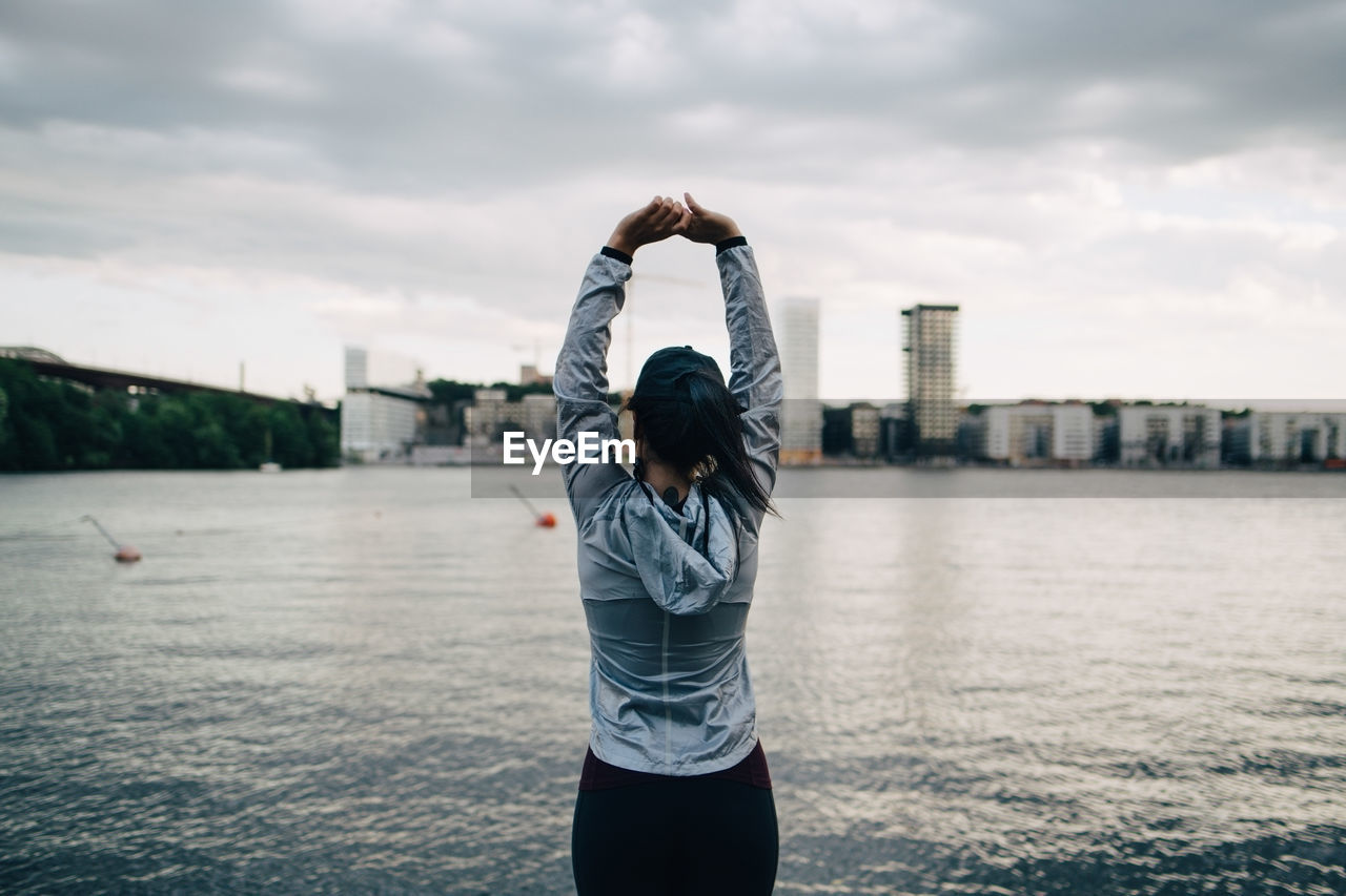 Rear view of female athlete stretching arms while standing against sea in city