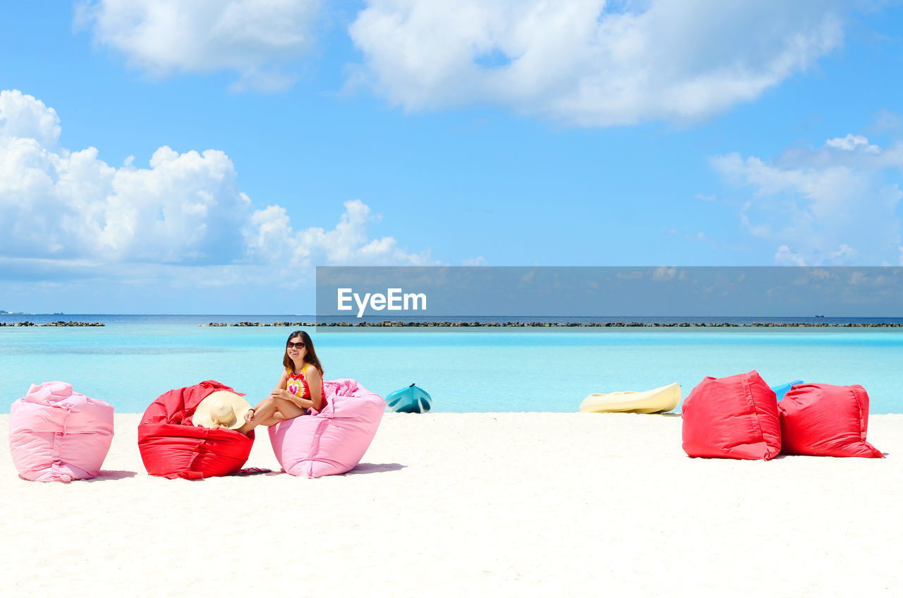 Woman resting on bean bags at beach against sky during sunny day