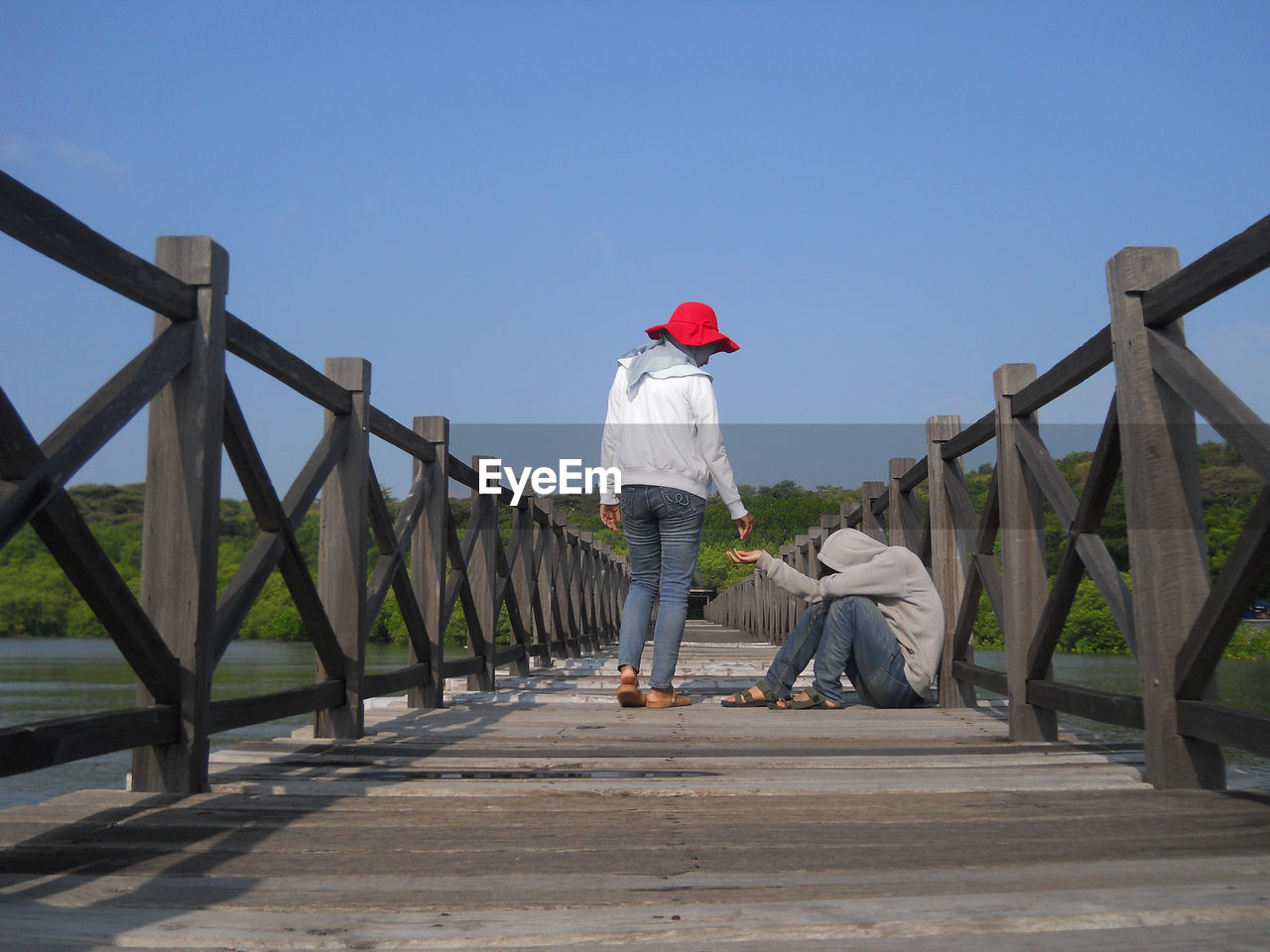 Rear view of woman giving money to beggar sitting on footbridge against clear sky