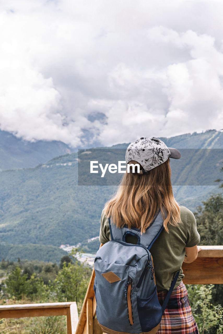 Rear view of young blonde woman in cap looking at view of mountains on wooden viewpoint, hiking 