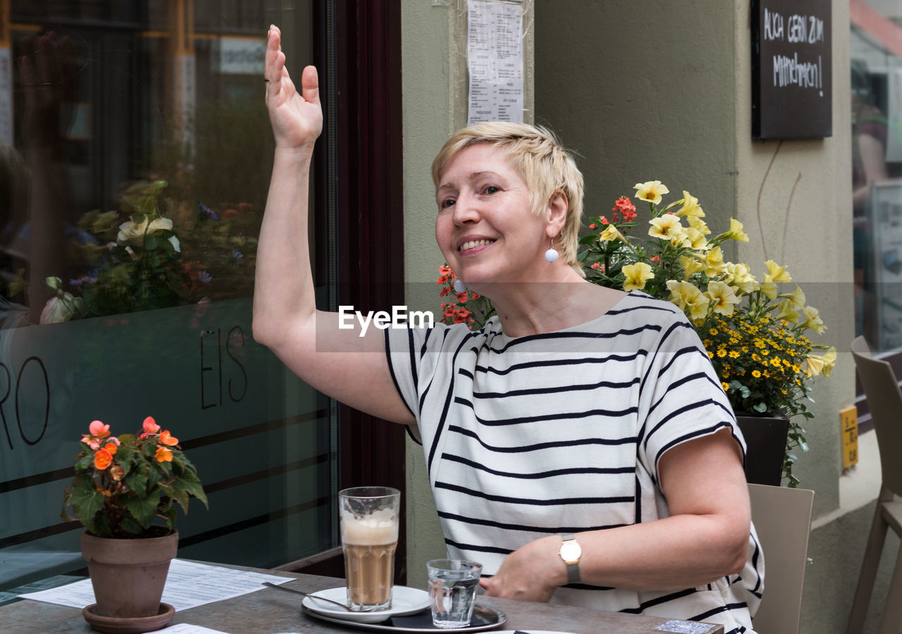 An attractive middle-aged woman sits in a street café in the summer and waves cheerfully