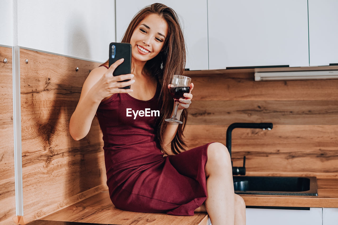 YOUNG WOMAN USING PHONE WHILE SITTING ON WOODEN FLOOR