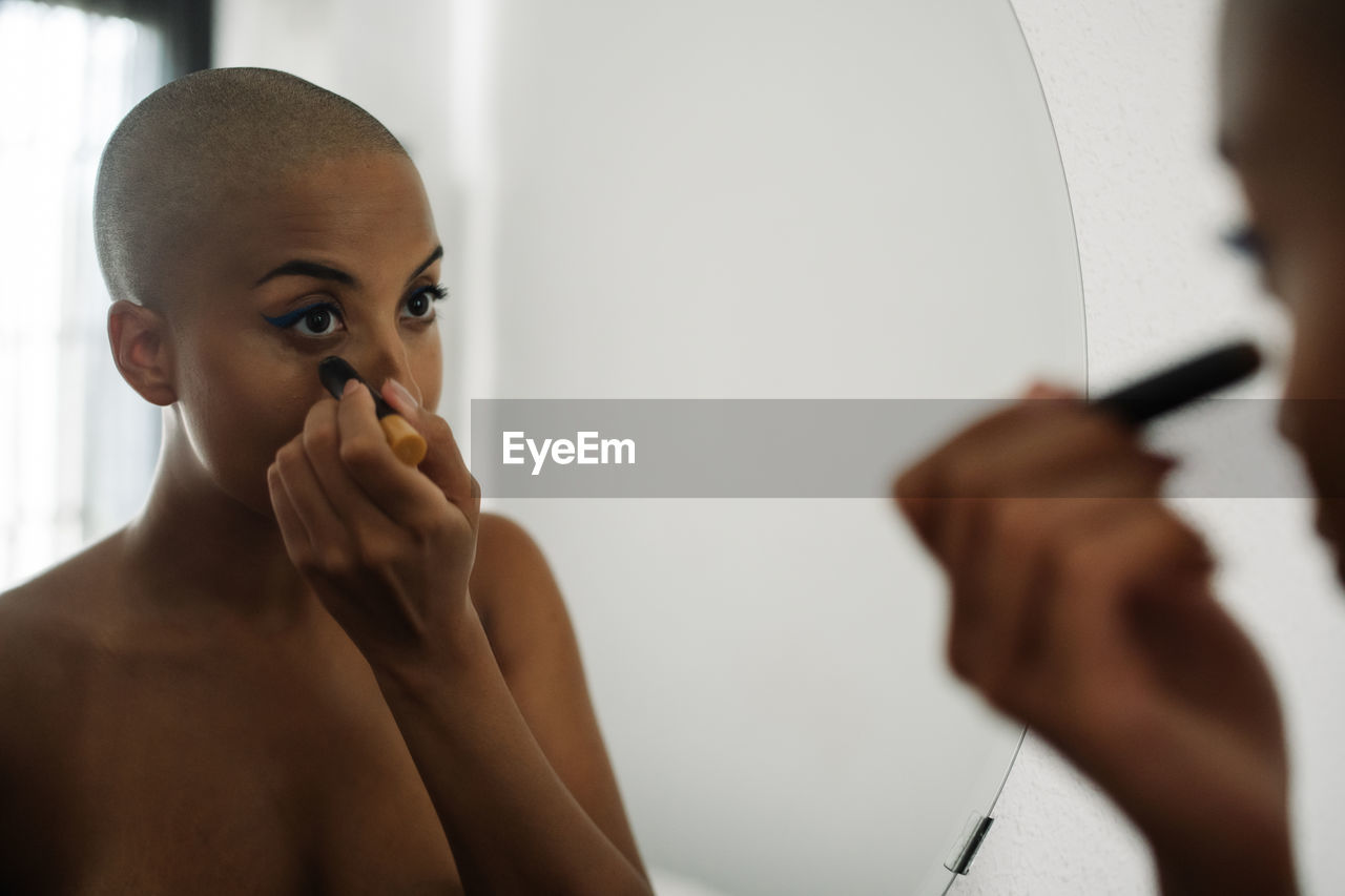 Concentrated african american female with bald haircut looking in mirror and applying foundation with brush while doing makeup