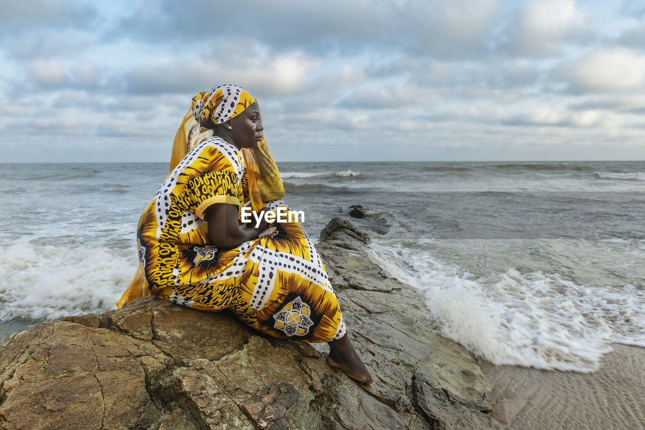 African woman with long dress sitting on stone cliff after the coast in accra ghana west africa