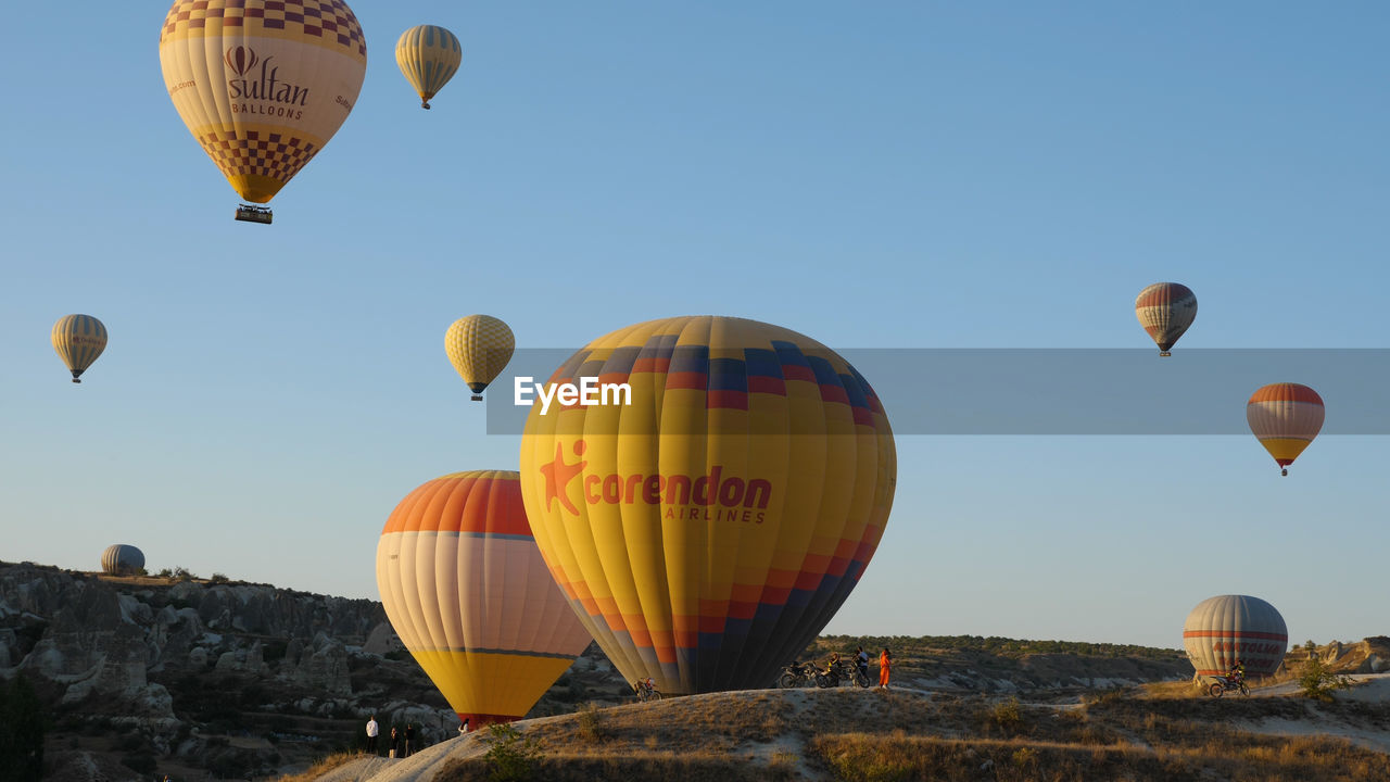 hot air balloon, balloon, hot air ballooning, vehicle, aircraft, air vehicle, transportation, mid-air, flying, adventure, ballooning festival, mode of transportation, basket, sky, travel, multi colored, nature, toy, rock, journey, taking off, environment, travel destinations, tourism, cloud, heat, morning, blue, clear sky, executive car, celebration, tradition, outdoors, landscape, large group of objects, container, day, recreation, rock hoodoo, trip, vacation, rock formation, traditional festival, mountain, group of objects, motion, holiday, no people, leisure activity, flame, burning, low angle view, outdoor pursuit, dawn