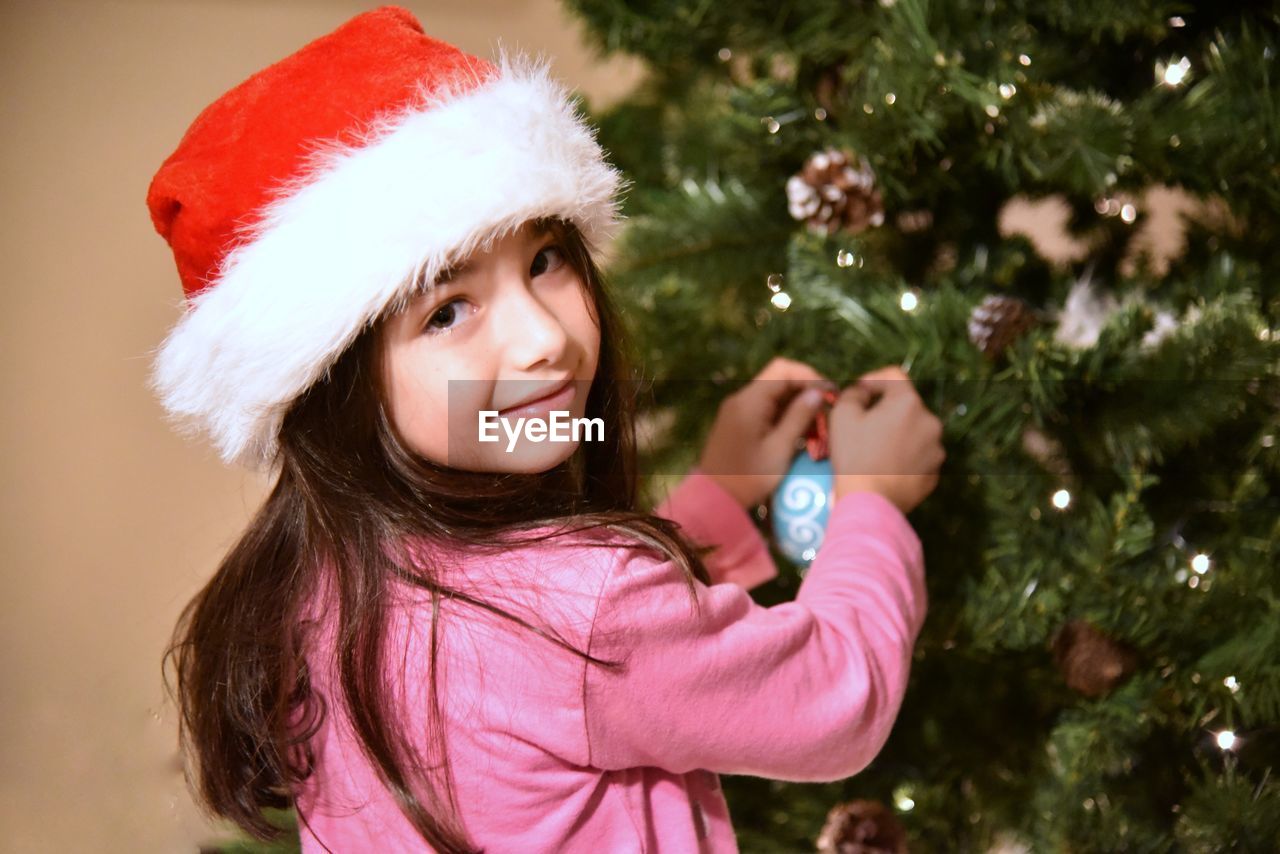 Portrait of smiling girl wearing santa hat while decorating christmas tree at home