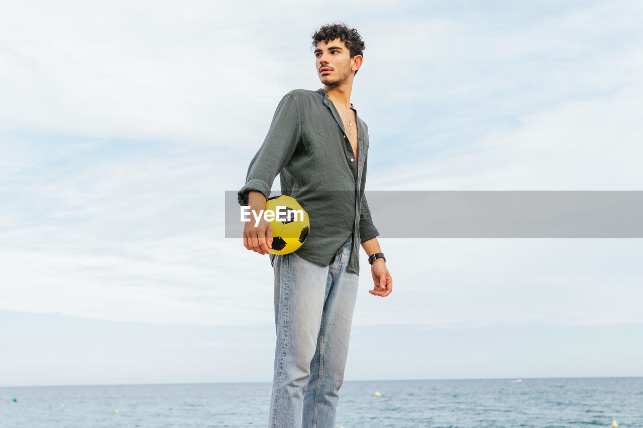 Hispanic male in shirt and jeans holding yellow football ball looking away while spending time on sandy beach near sea