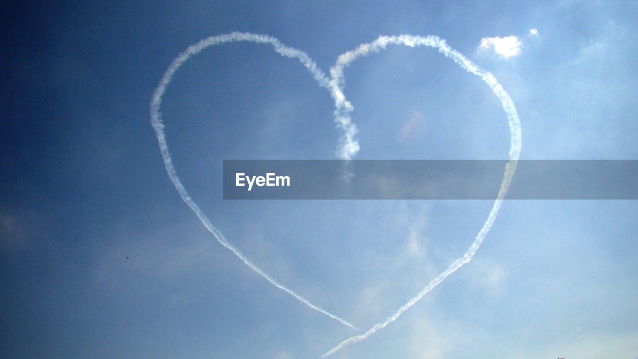 Low angle view of heart shape vapor trail in blue sky