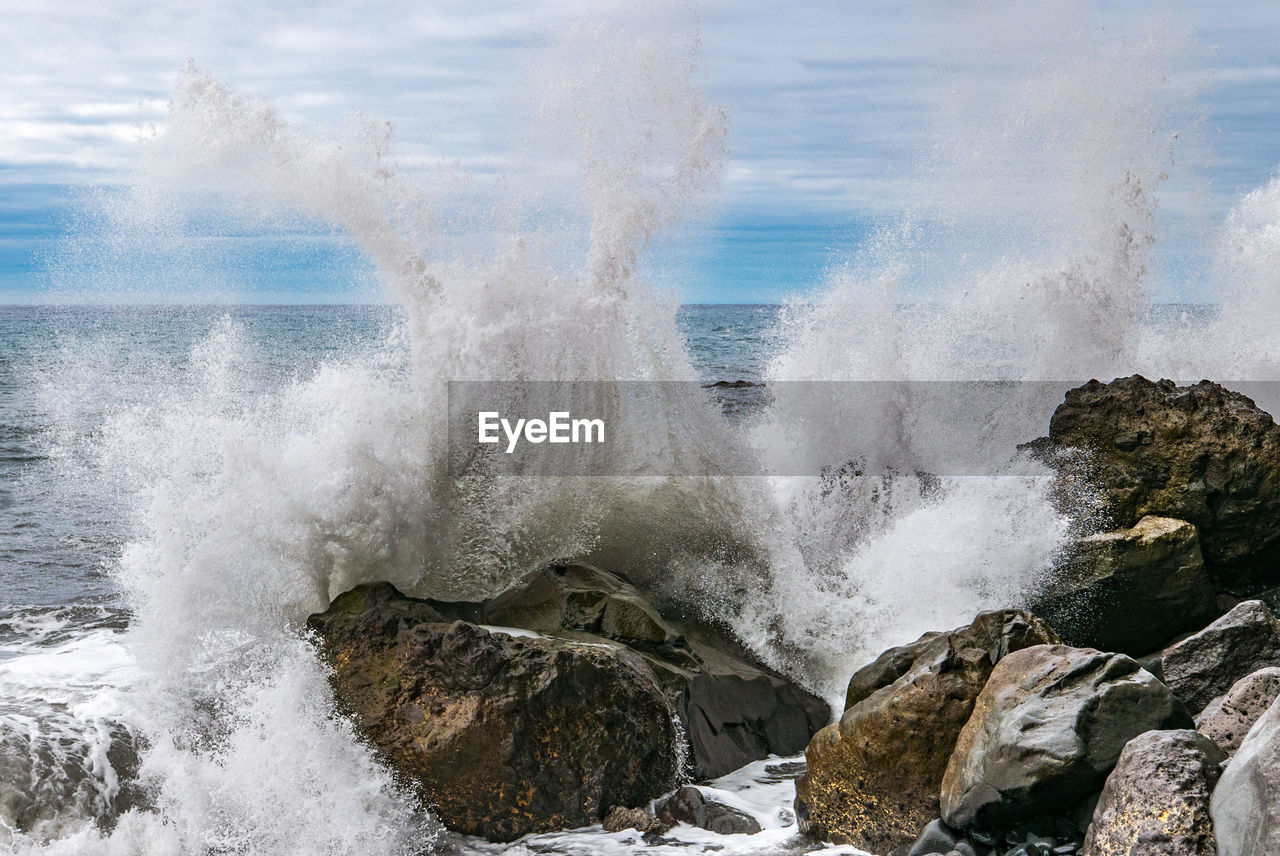 Waves splashing on rocks at shore against cloudy sky