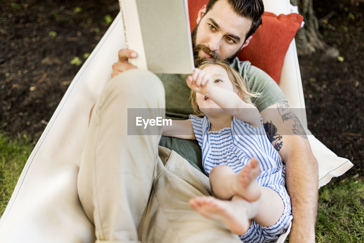 High angle view of daughter with father reading book while lying on hammock at yard