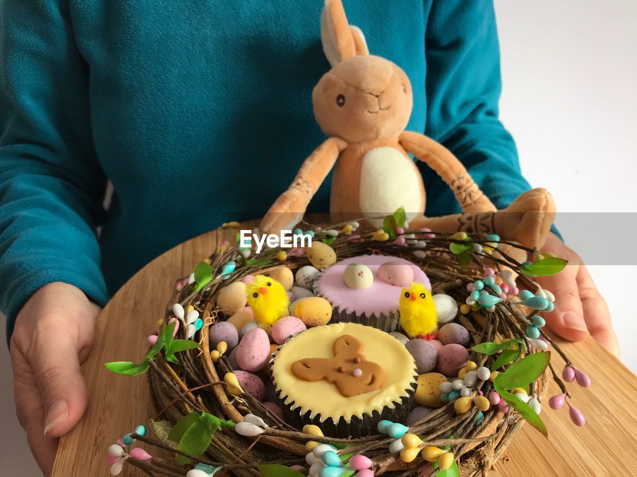 Woman holding  bamboo board with iced cupcakes and chocolate easter eggs in a decorative nest shape.