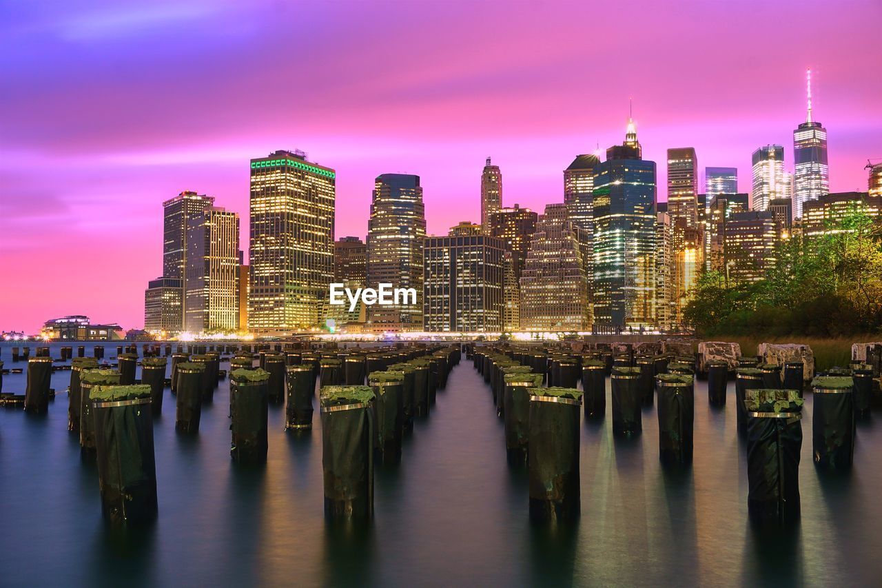 Wooden posts in river against modern buildings at night