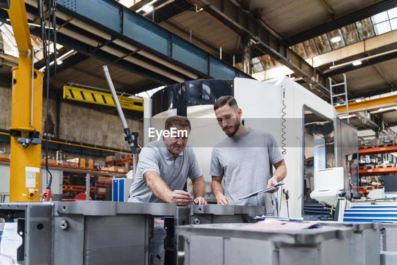 Male engineer examining product standing by young coworker in industry