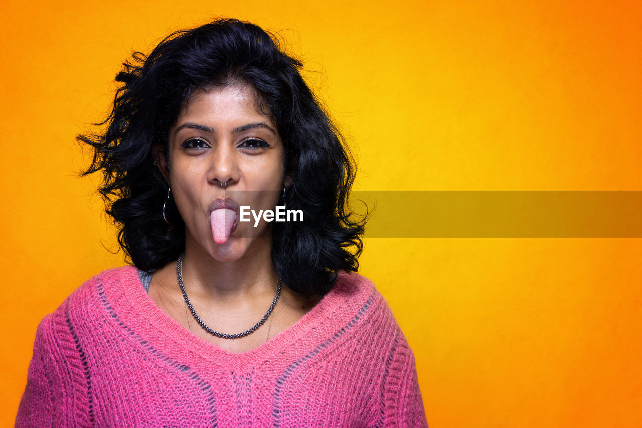 Portrait of young woman sticking out tongue against orange background