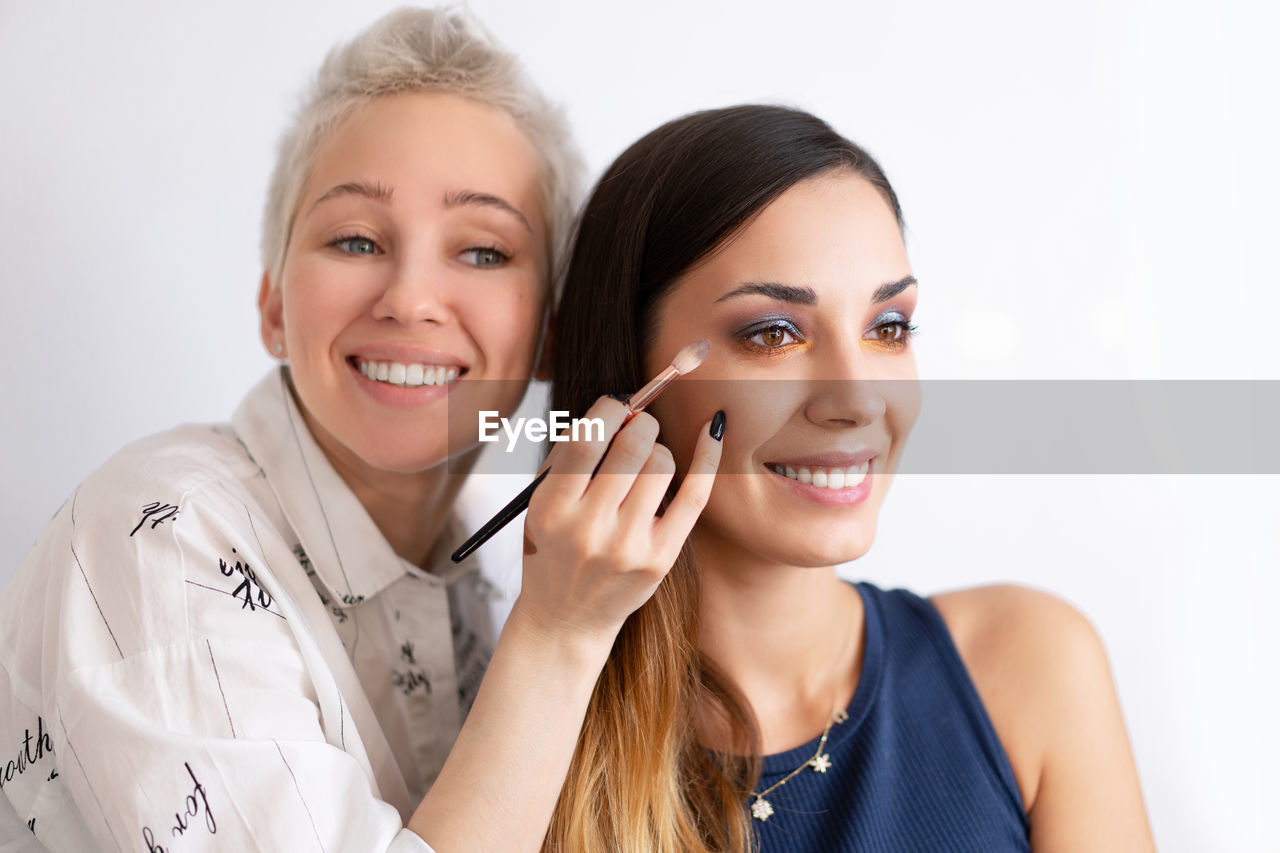 Smiling beautician applying make-up to fashion model against white background