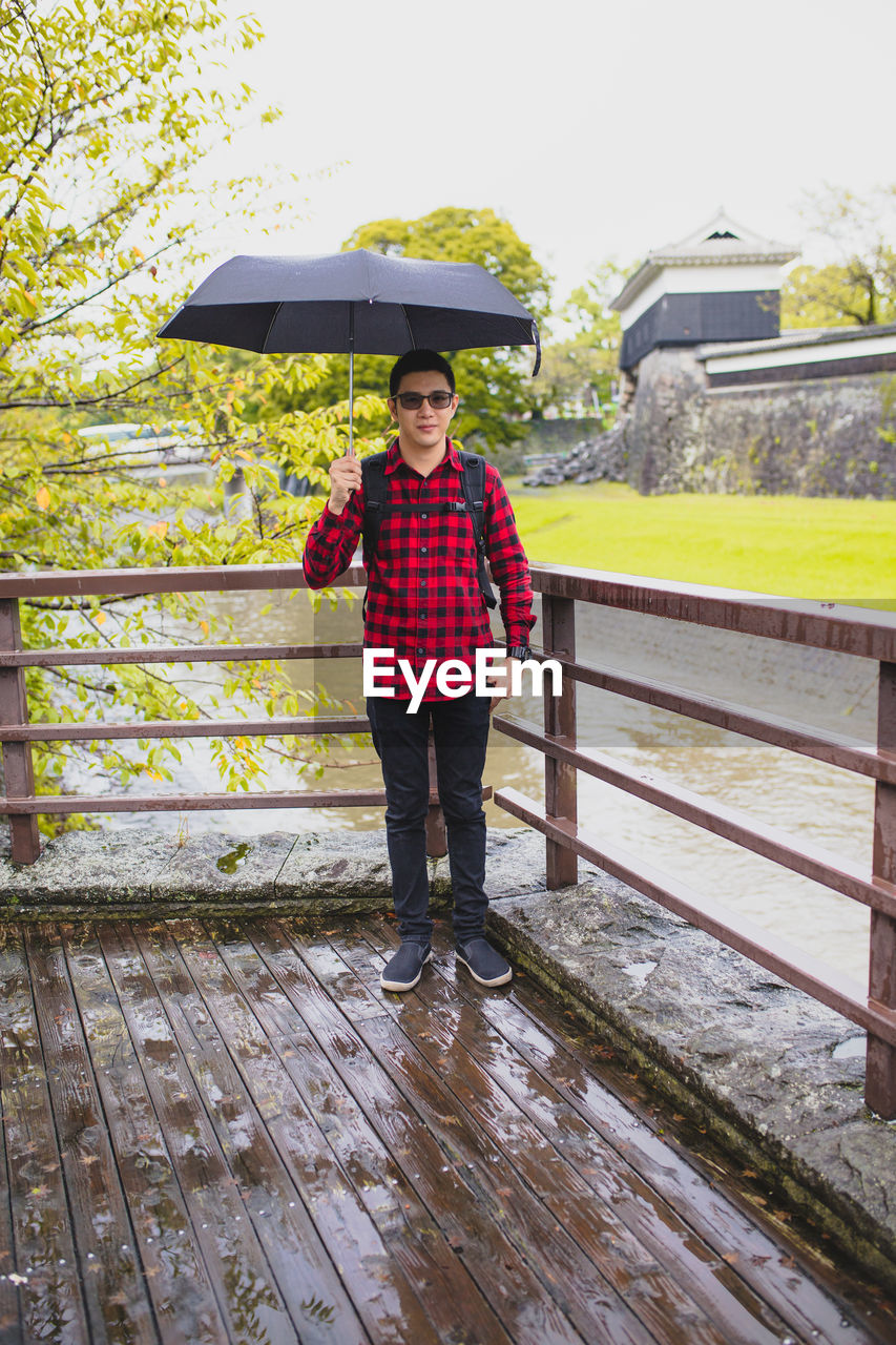 Portrait of man with umbrella standing on bridge during rainy season