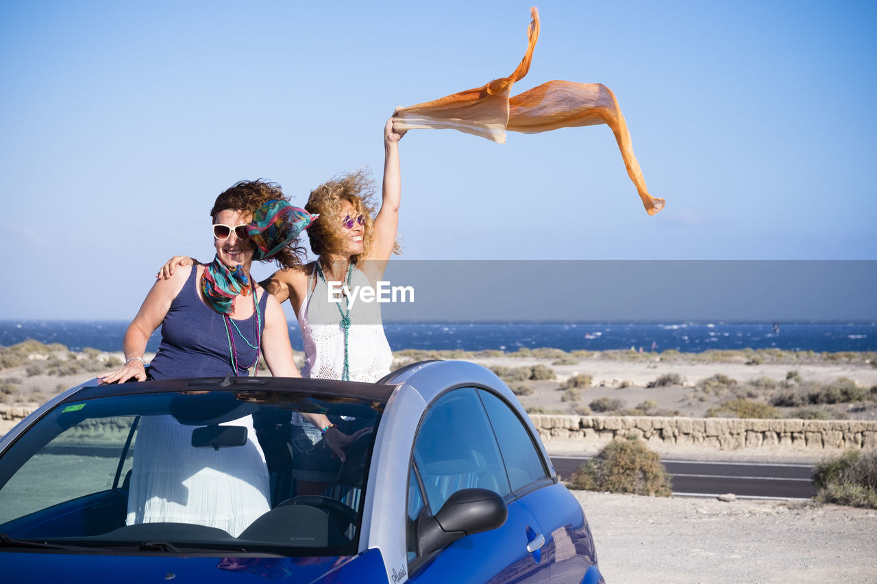 Happy female friends standing in car at beach against clear blue sky