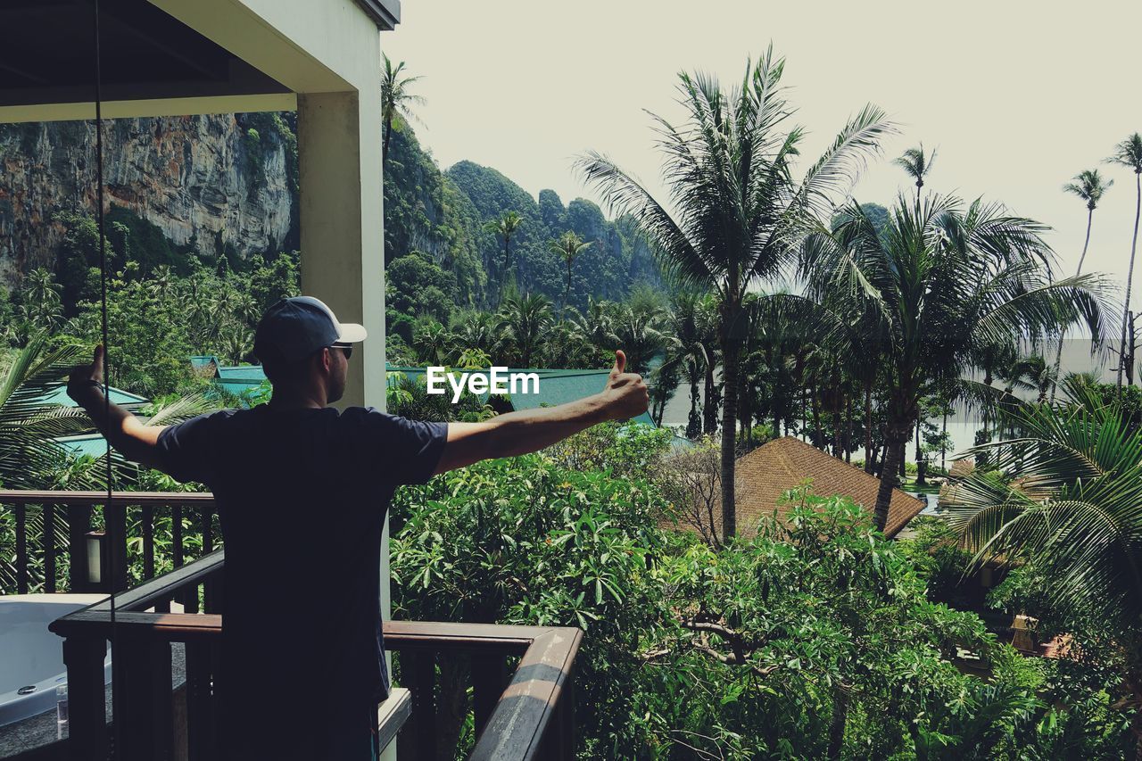 Rear view of man gesturing thumbs up while standing at balcony against trees and mountains