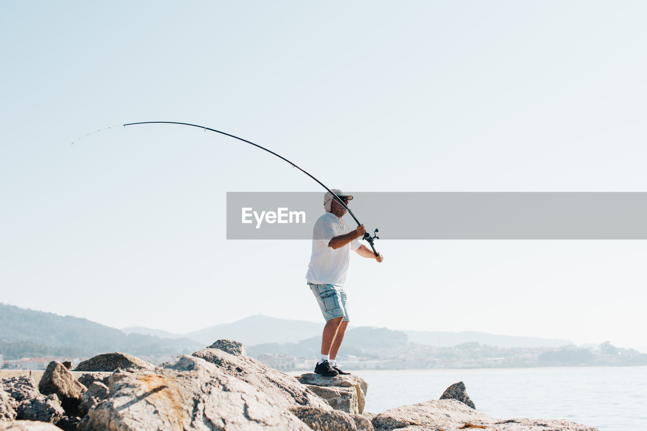 Side view of enthusiastic man in cap fishing and throwing rod into sea water standing on stony shore in bright day