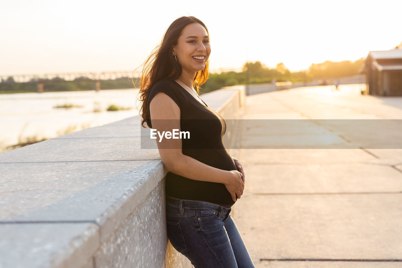 PORTRAIT OF SMILING YOUNG WOMAN STANDING AGAINST THE SKY