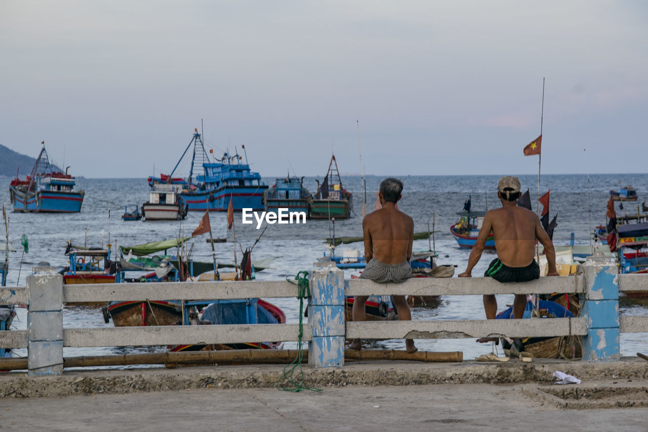 REAR VIEW OF PEOPLE SITTING ON BEACH AGAINST SKY