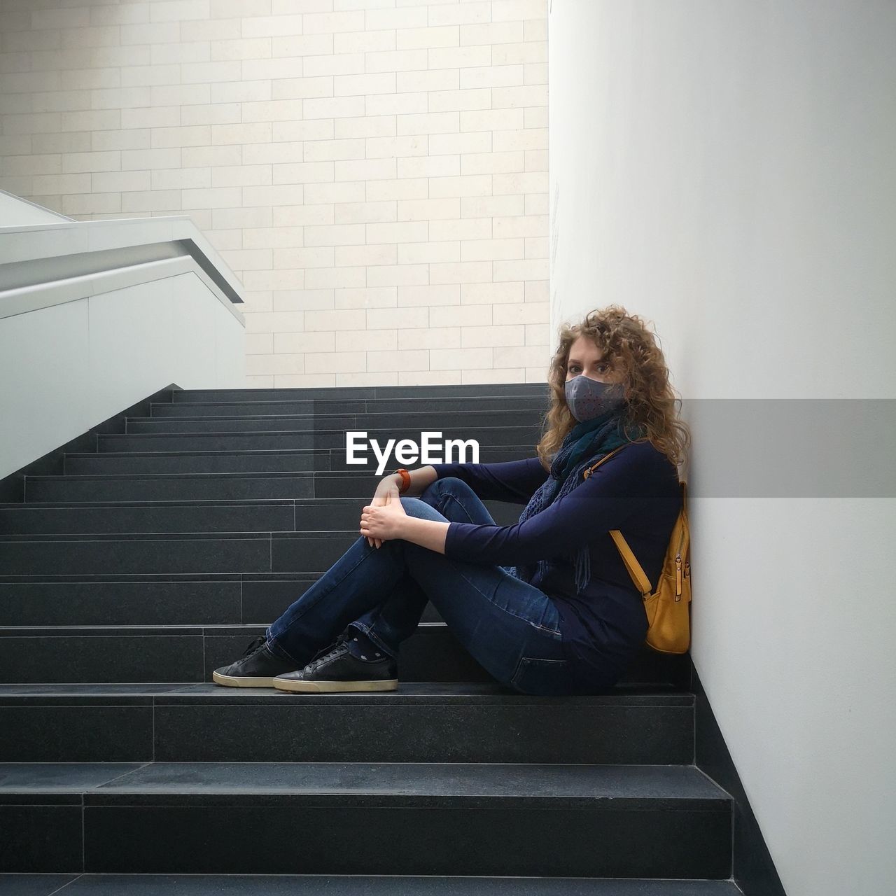 Young curly woman in a protective mask and jeans sits on the stairs of an empty museum building