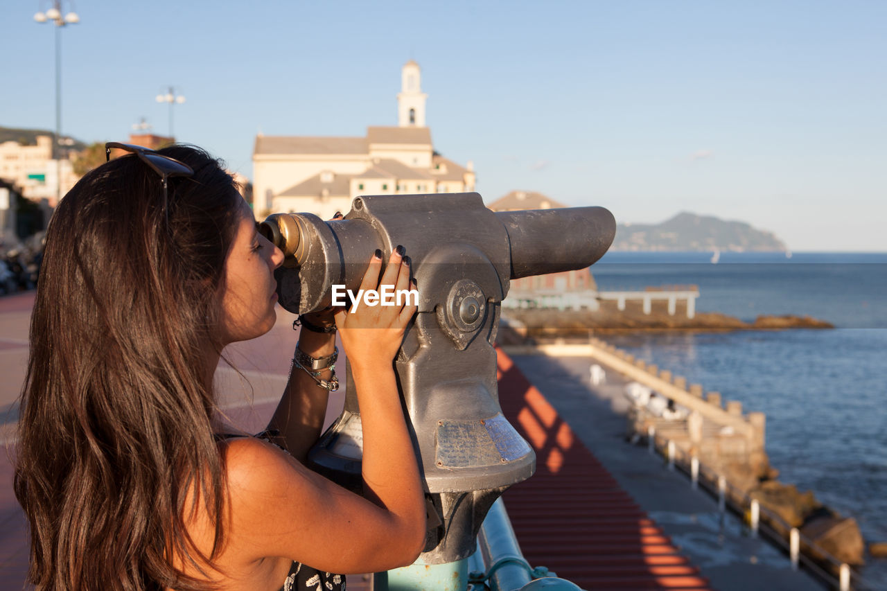 Side view of young woman looking at sea through coin-operated binoculars in city