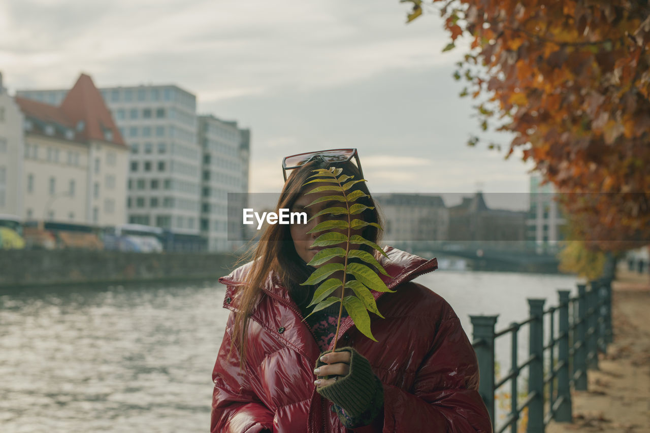 Portrait of woman holding leaf against face while standing by railing in city