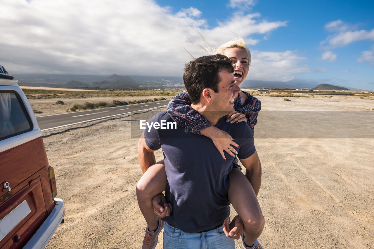 Happy man carrying woman while walking at beach against sky