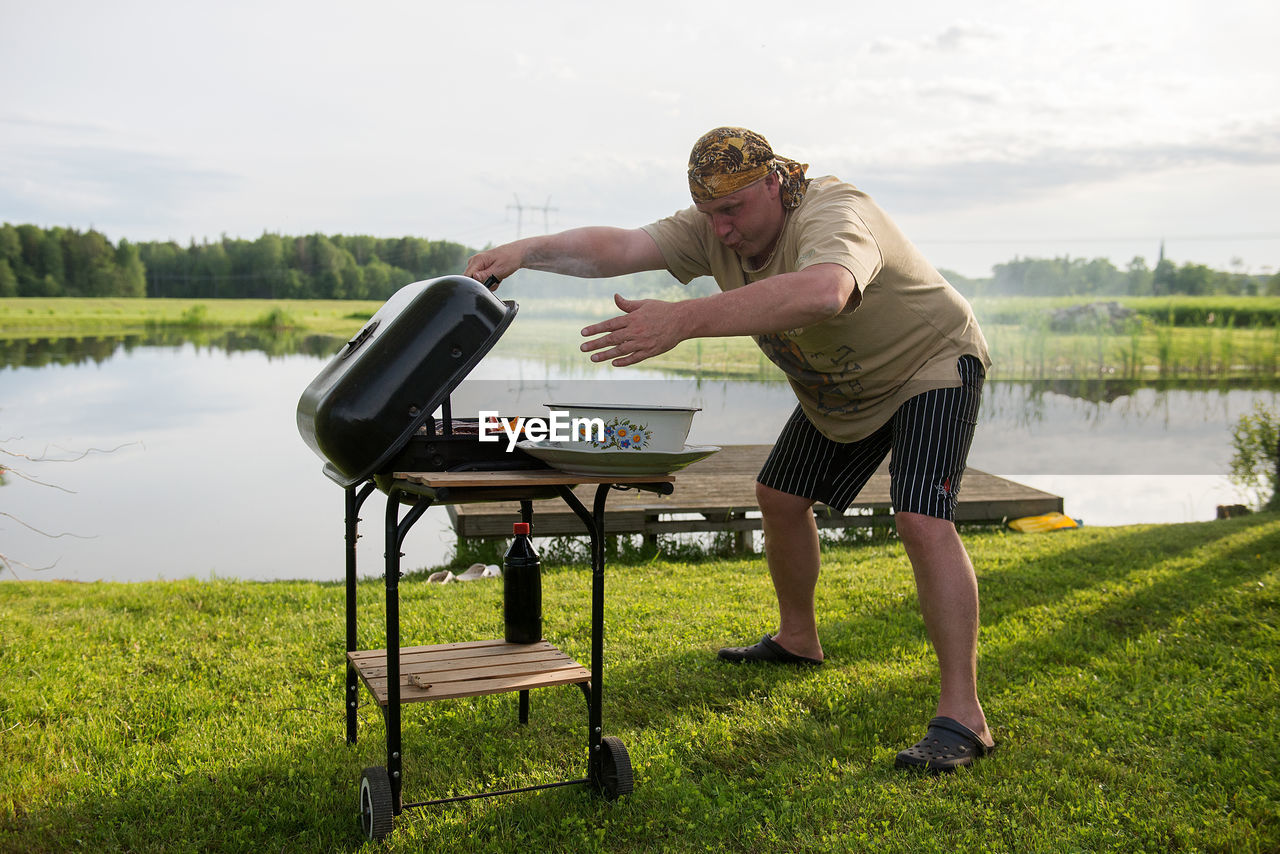 Young man grilling by lake against sky