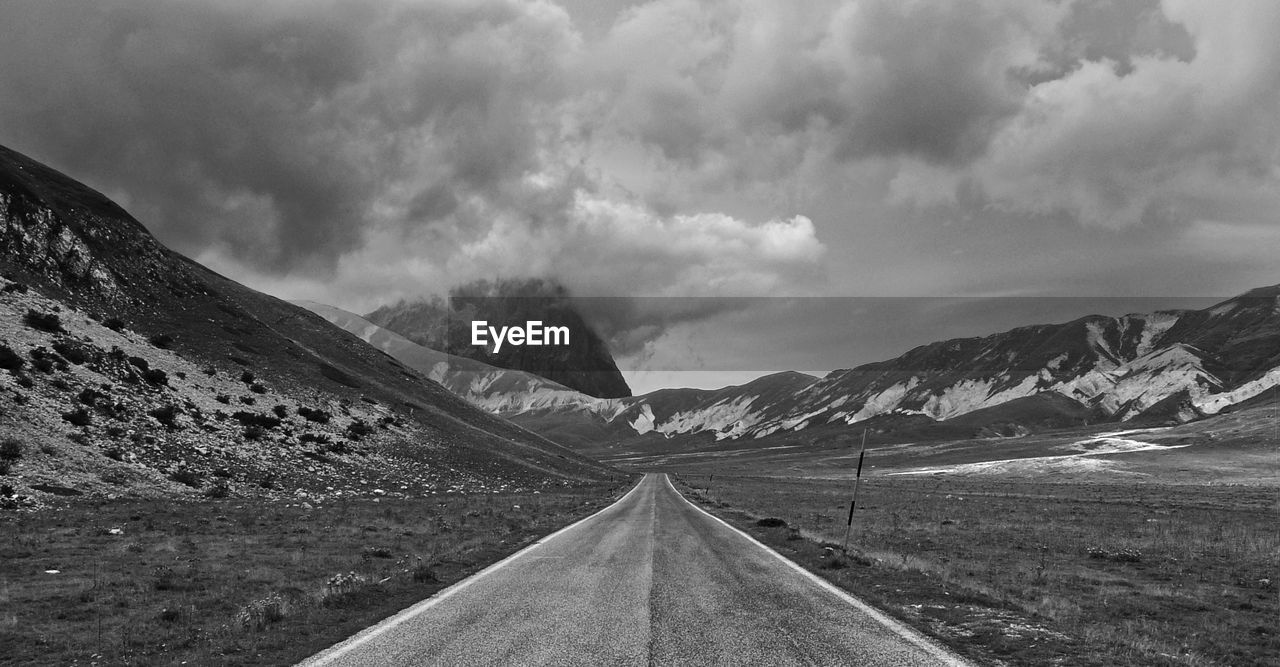 Empty road on landscape by mountains against cloudy sky