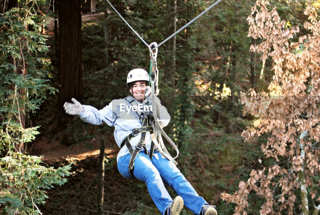 Portrait of smiling woman hanging from zip line against trees in forest