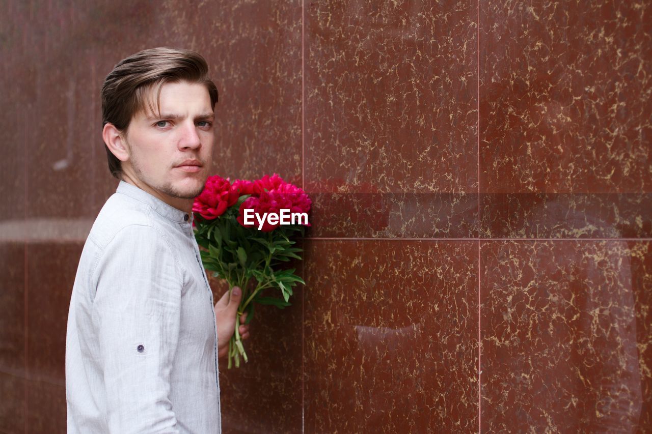 Side view of handsome young man with flowers standing by wall